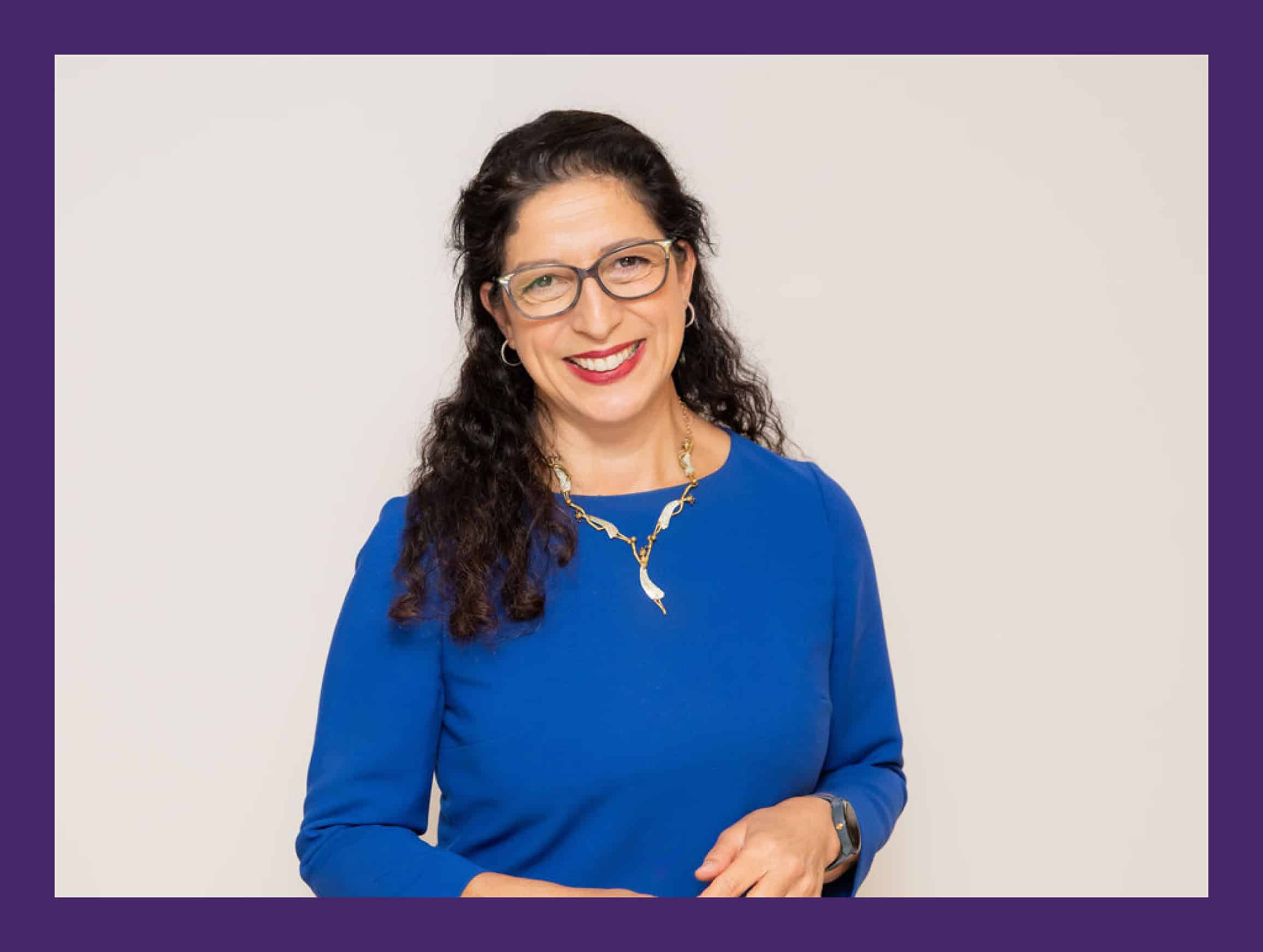 A woman with long dark hair wearing glasses and a blue top smiles at the camera